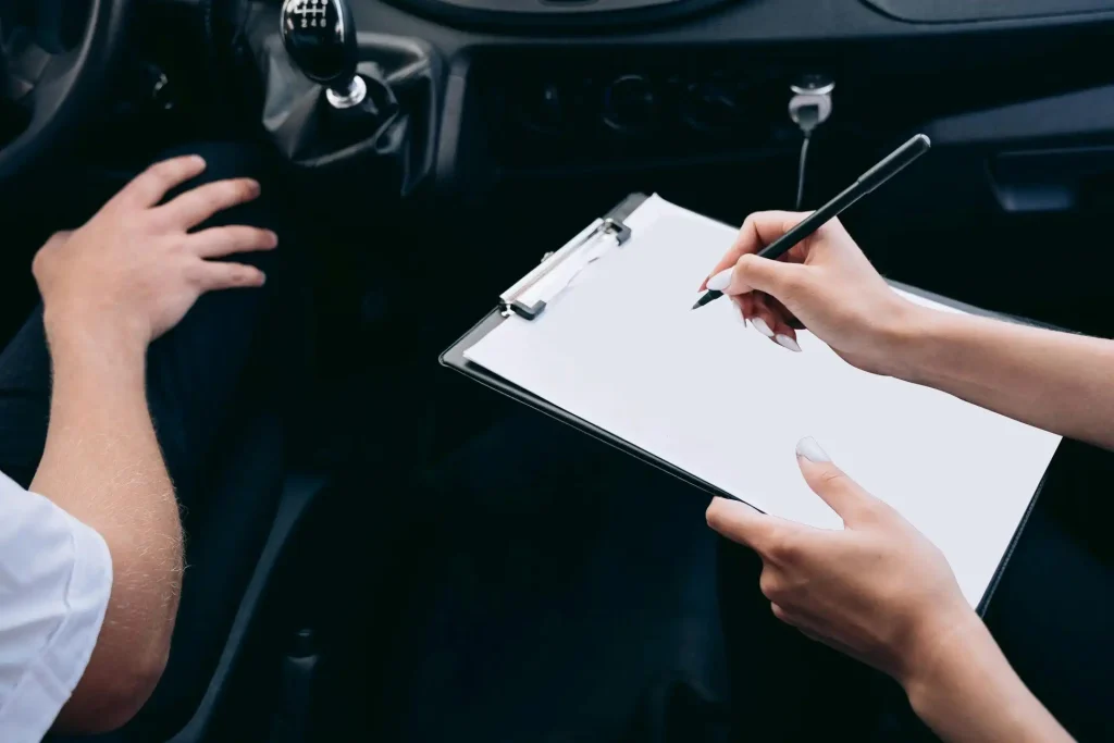 Nahaufnahme einer Person, die in einem Auto auf einem Klemmbrett schreibt; Hände und Schalthebel sind zu sehen.